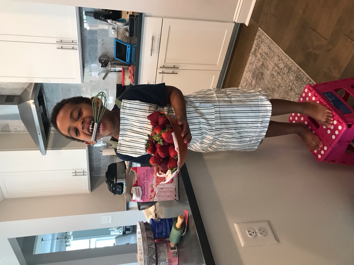Yonas grinning in the kitchen wearing a striped apron, holding a bowl of fresh strawberries and a whisk