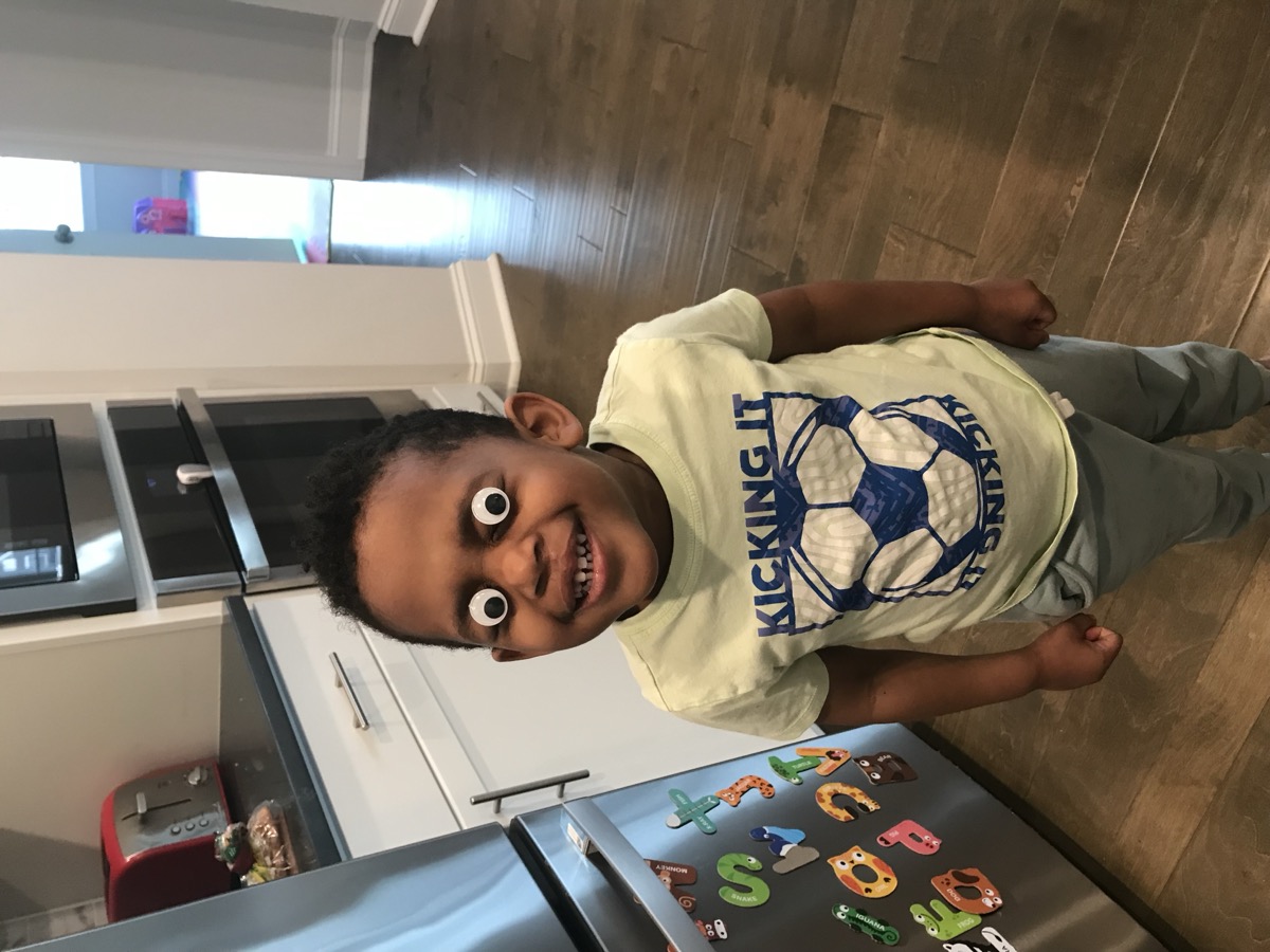 Yonas smiling big with googly eyes on his face, wearing a Kicking It soccer shirt in the kitchen