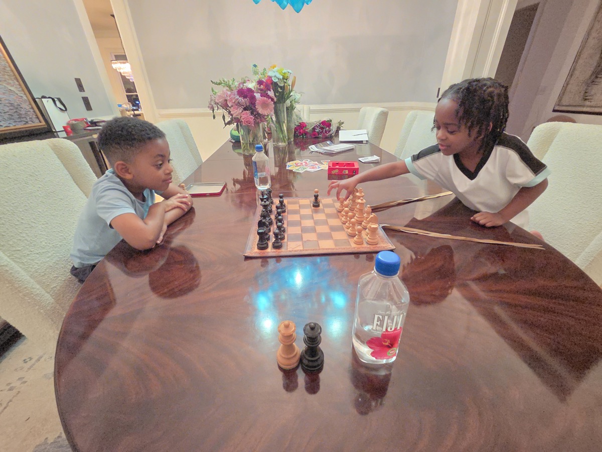 Yonas playing chess at the dining table with his twin sister, studying the board intently in a light blue shirt