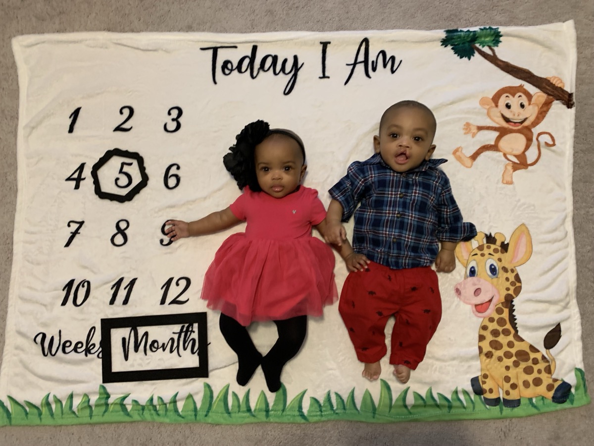 Yonas and his twin on a milestone blanket marking 5 months, Yonas in a navy plaid shirt and red pants, holding hands with his sister