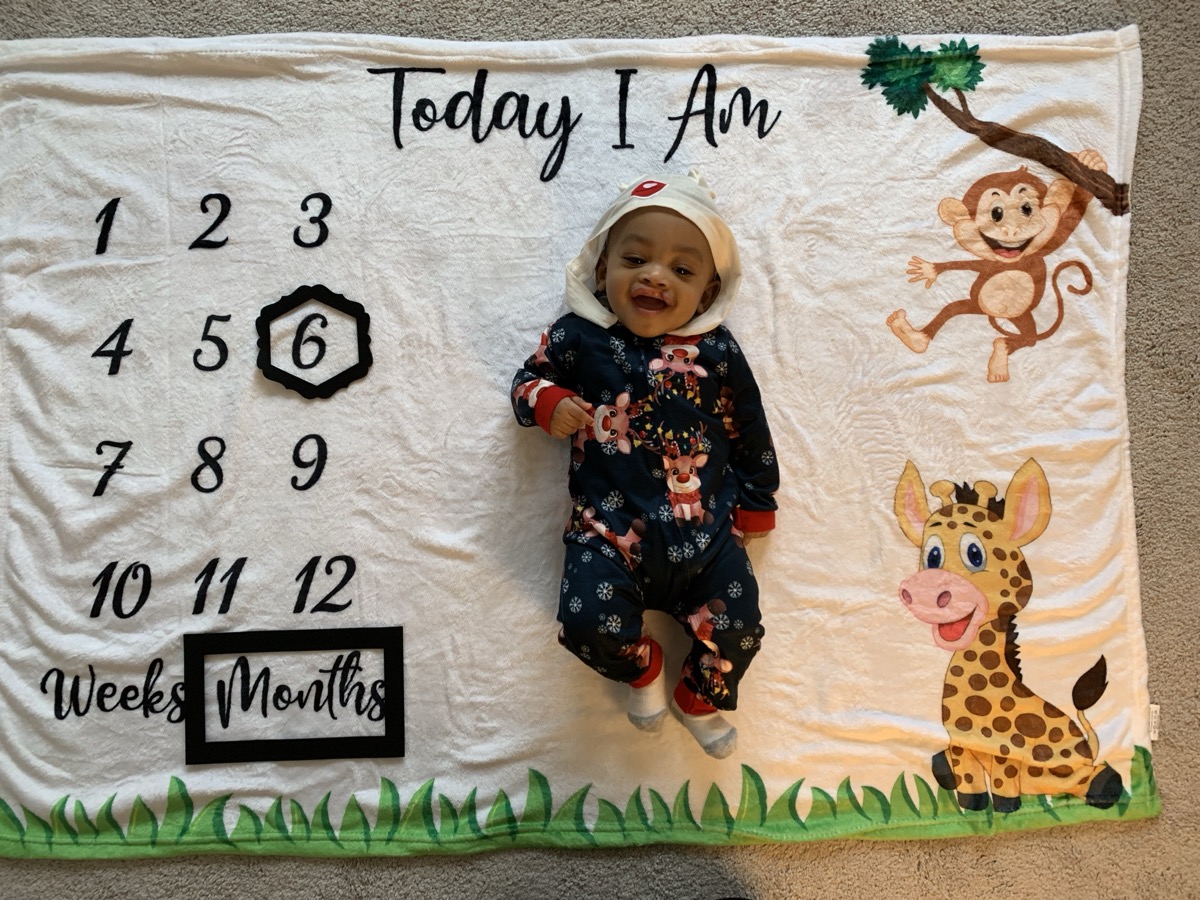 Baby Yonas smiling big on a milestone blanket marking 6 months, wearing a festive holiday reindeer onesie with hood