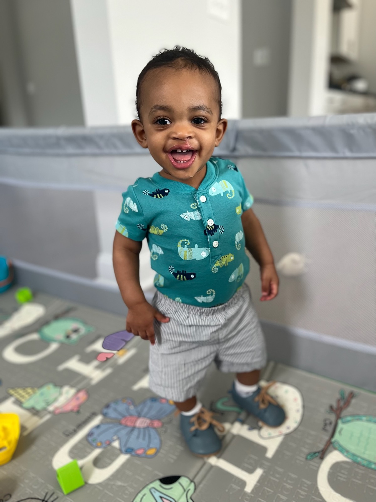 Toddler Yonas standing and grinning in a playpen, wearing a teal chameleon-print henley shirt and gray striped shorts