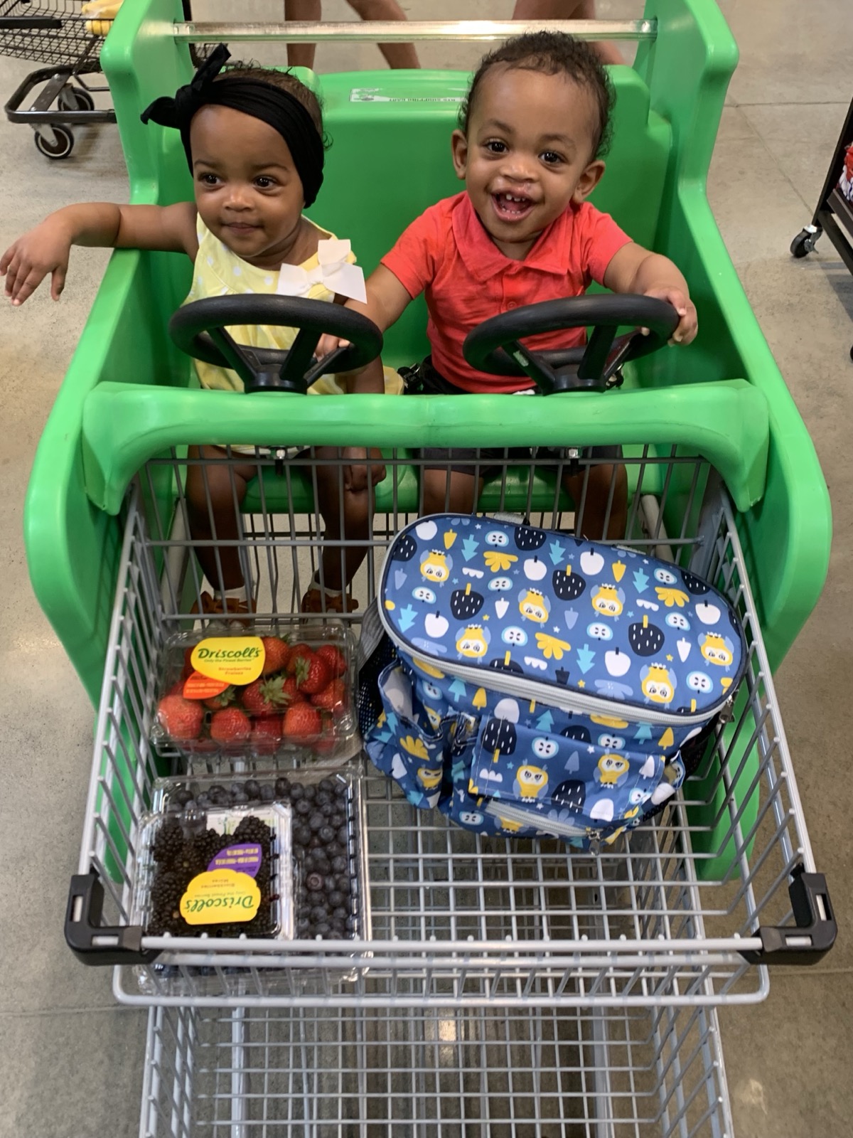 Yonas and his twin sister riding in a green grocery cart together, Yonas in a red polo shirt with fresh berries in the cart