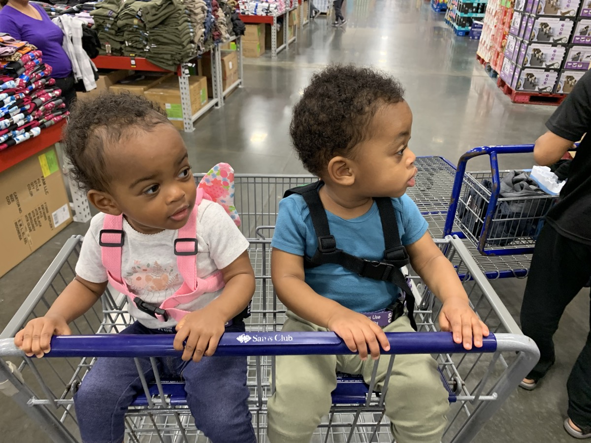 Yonas and his twin sister sitting side by side in a Sam's Club shopping cart, Yonas in a blue shirt looking to the side with curly hair