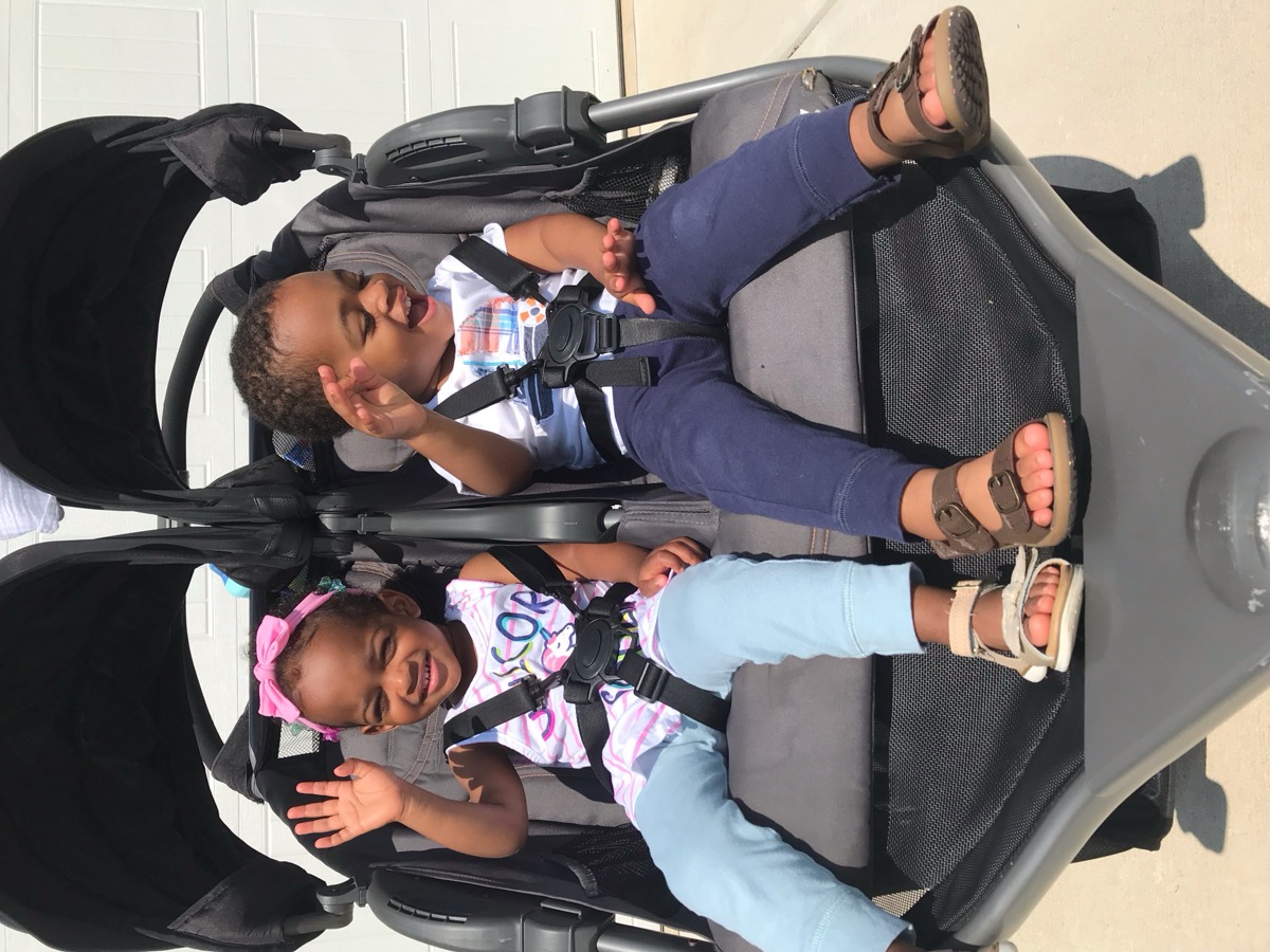 Yonas and his twin sister waving from a double stroller in the driveway, Yonas laughing in a white graphic tee and navy pants with sandals