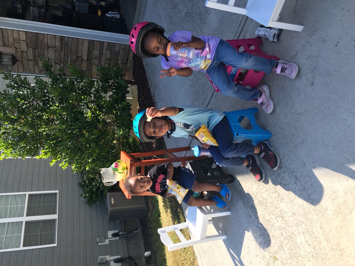 Yonas sitting on the driveway with two friends having snacks, wearing a Spider-Man shirt and blue sandals with a lemonade stand behind them