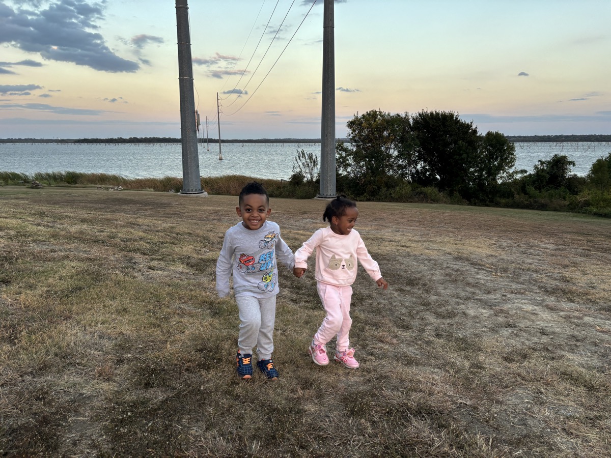 Yonas and his twin sister running together in a grassy field by a lake at sunset, Yonas smiling in a gray car-print sweatsuit