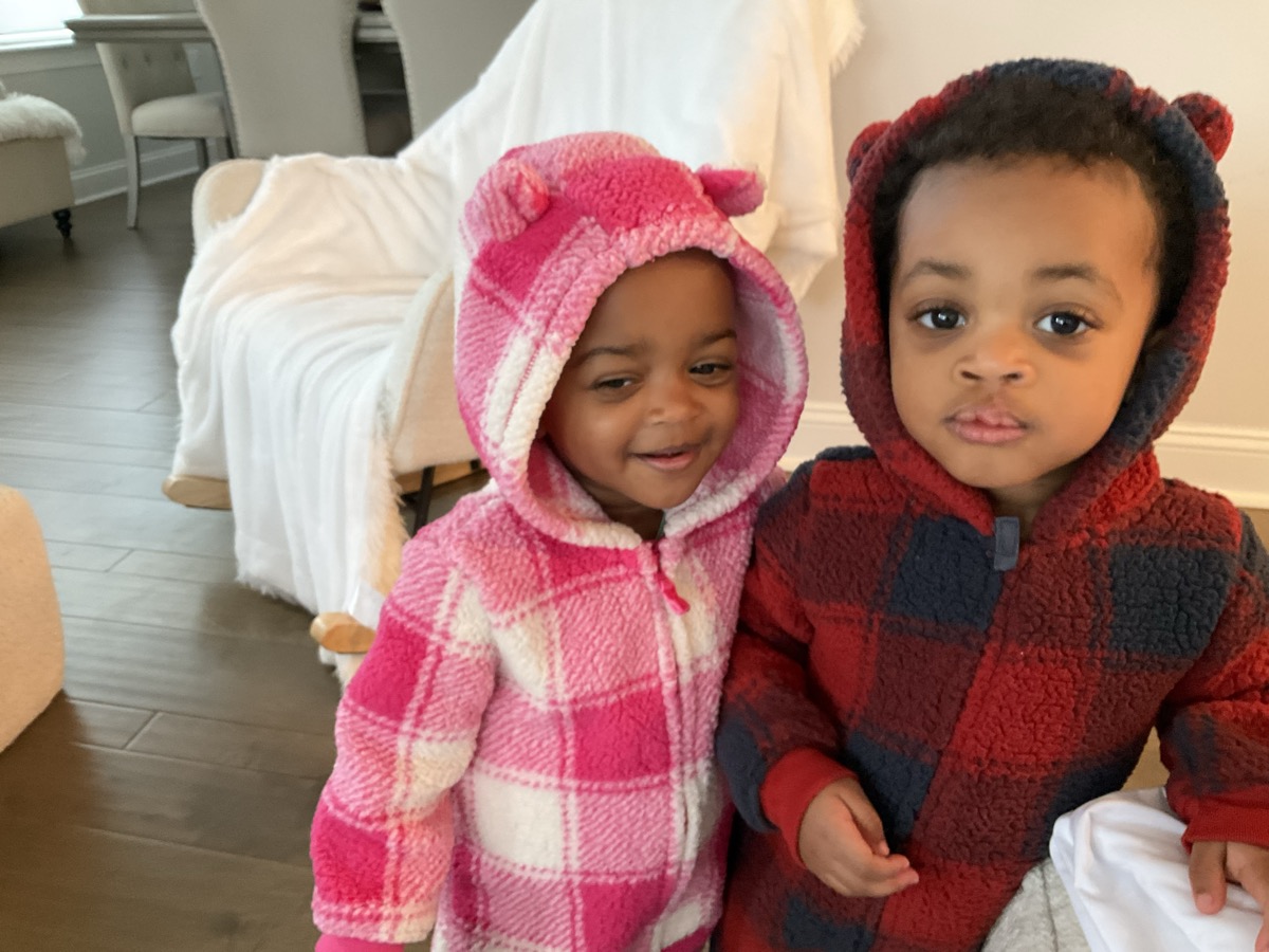 Yonas and his twin sister in cozy fleece hoodies with bear ears, Yonas in red and navy buffalo plaid, standing together indoors