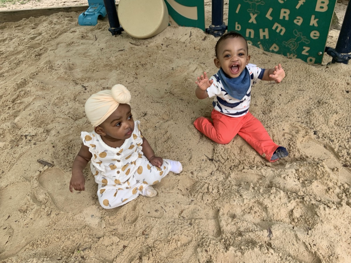 Baby Yonas laughing joyfully in a playground sandbox with his hands up, wearing a patterned shirt, blue bandana bib, and red pants