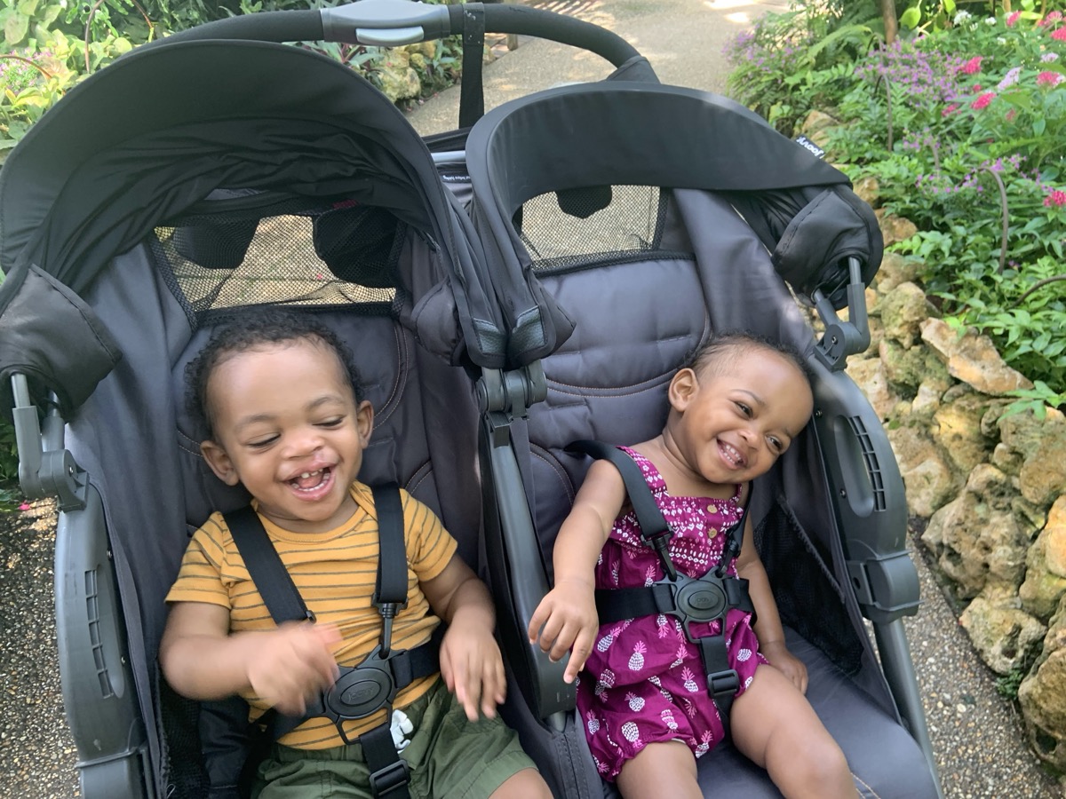 Yonas and his twin sister laughing together in a double stroller surrounded by lush garden greenery, Yonas in a yellow striped shirt