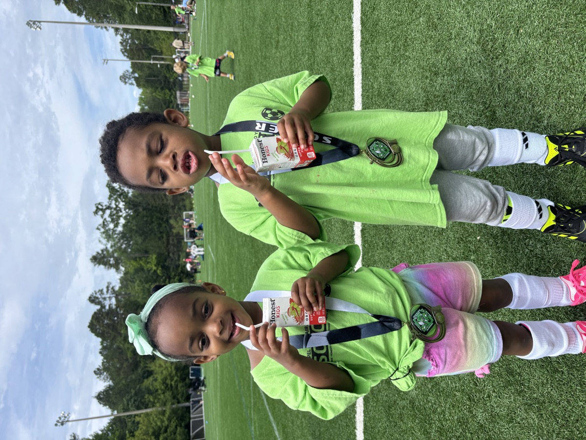 Yonas and his twin sister on a soccer field wearing matching green soccer jerseys with medals around their necks, sipping juice boxes