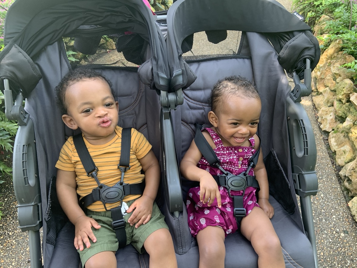 Yonas making a silly face in a double stroller at a garden, wearing a mustard striped tee and olive shorts, sitting next to his sister