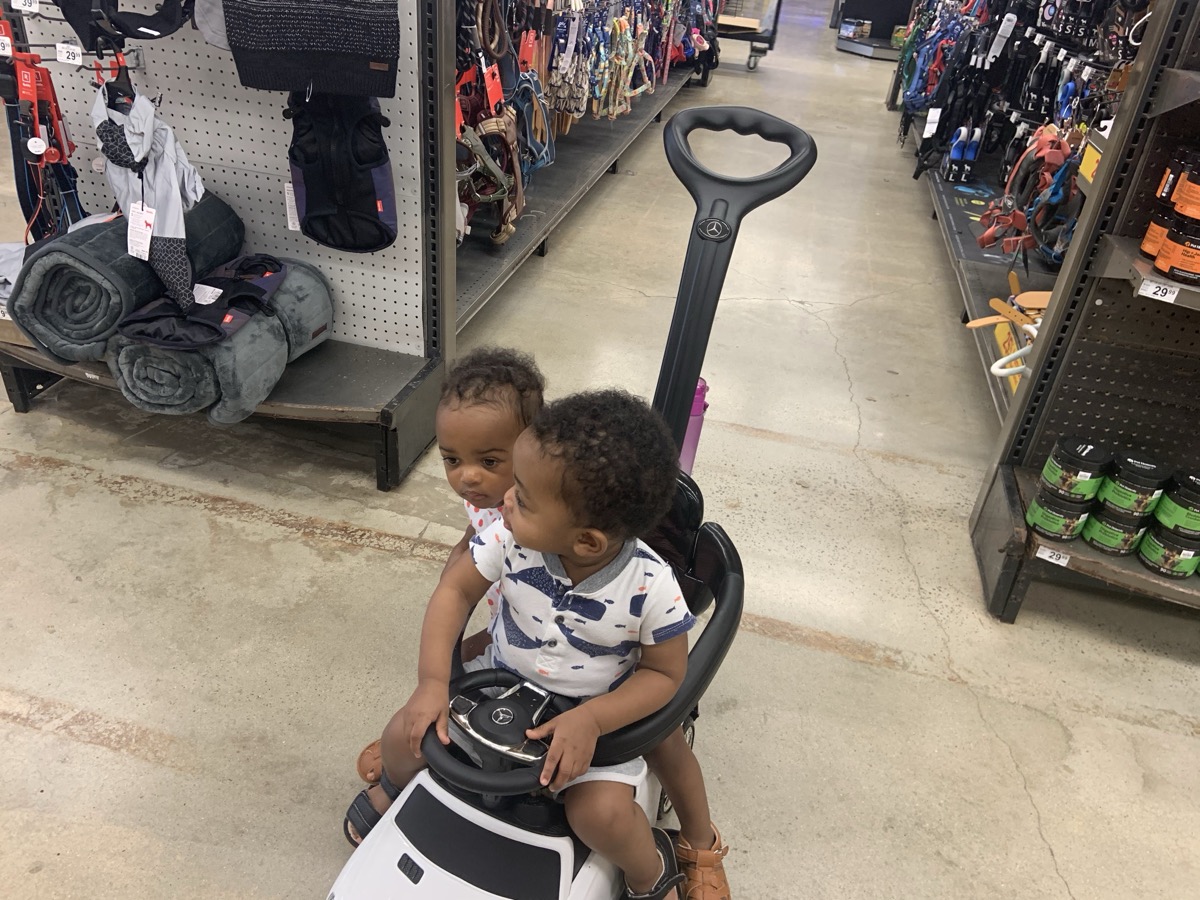 Yonas and his twin sister riding together on a small Mercedes push car in a store aisle, Yonas gripping the steering wheel