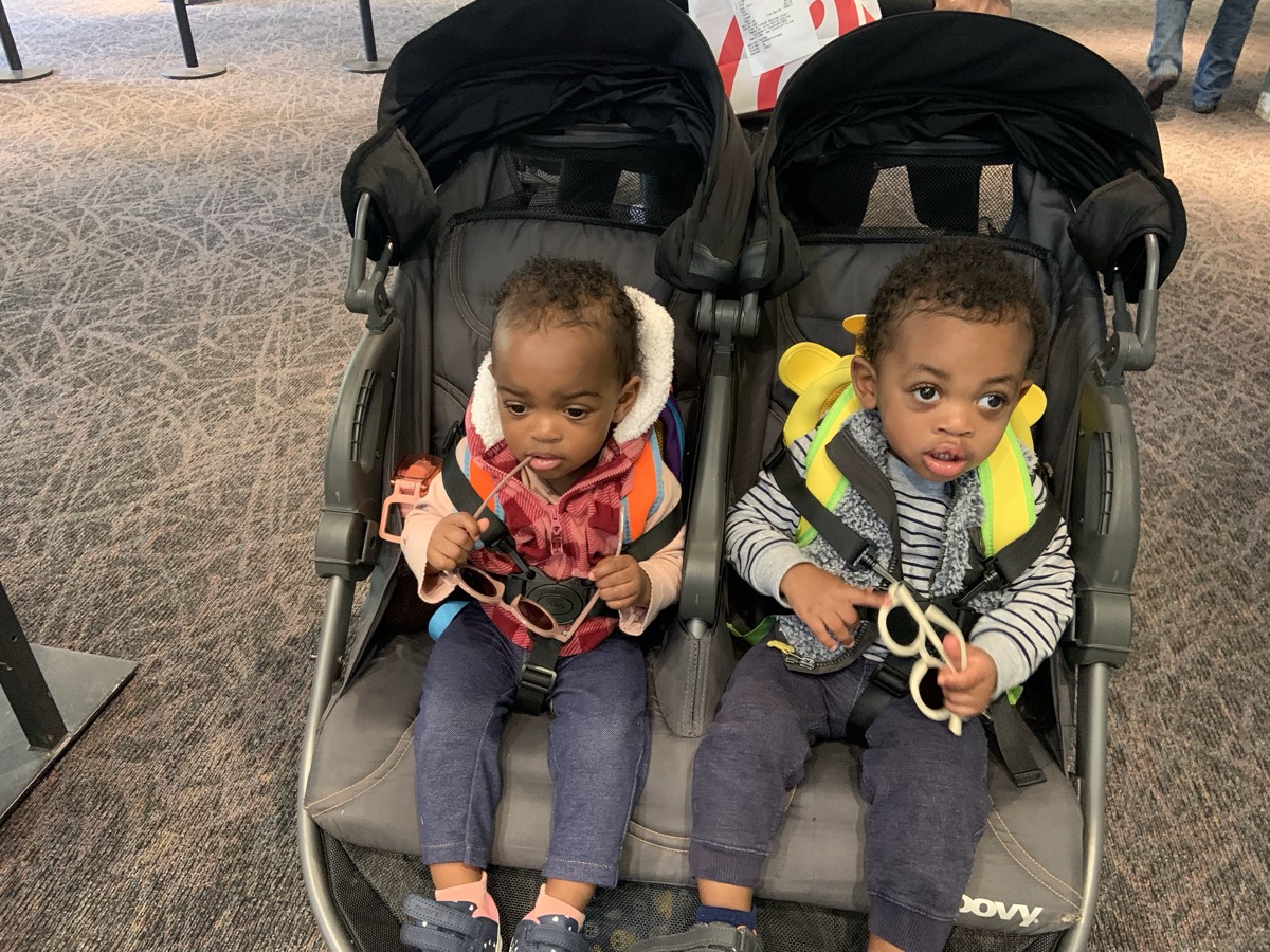 Yonas and his twin sister in a double stroller at an airport, each holding a pair of sunglasses, Yonas in a striped hoodie with layered jacket