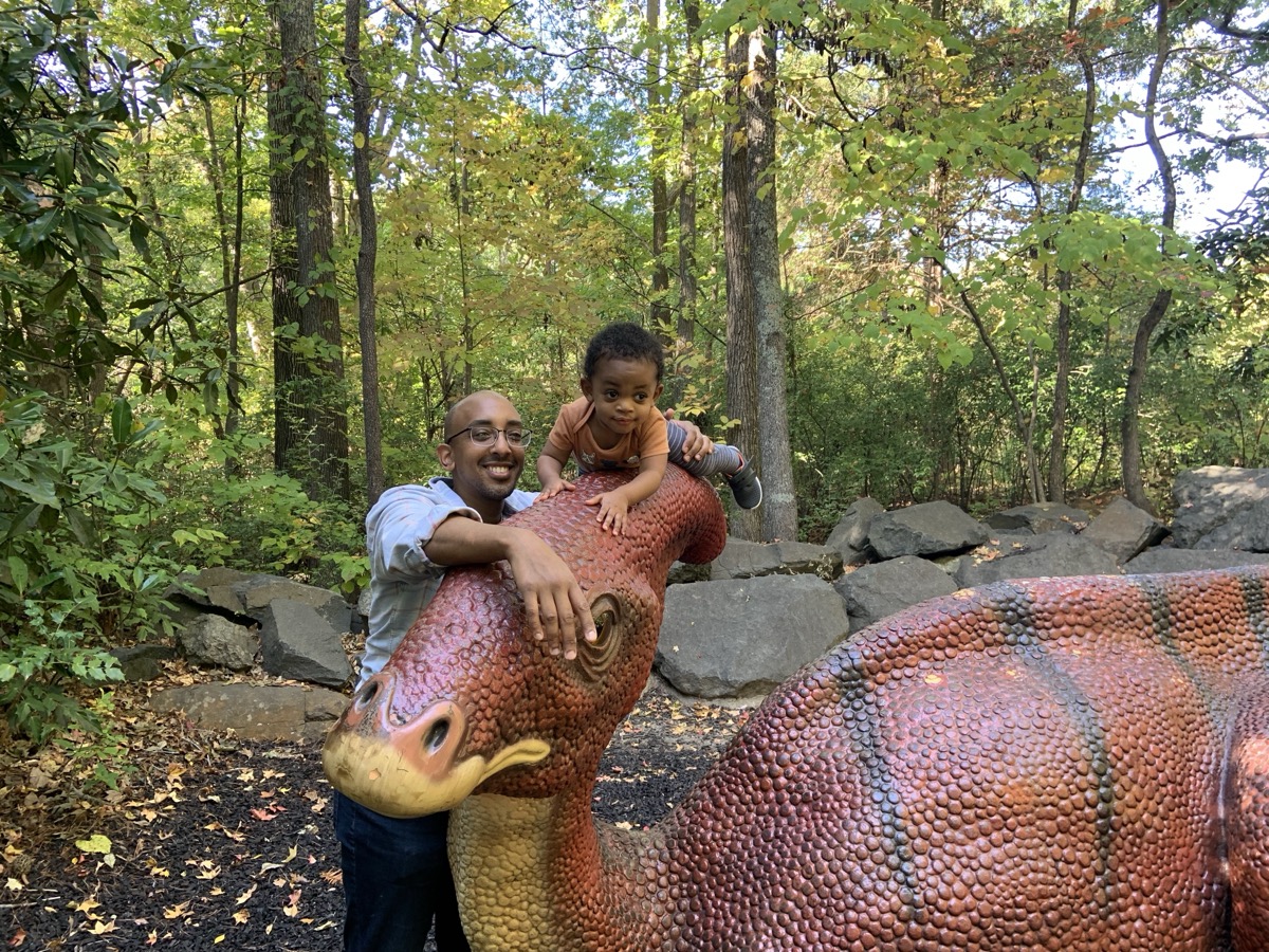 Yonas sitting on a dinosaur statue at a wooded park with his dad smiling behind him, Yonas in an orange-striped shirt climbing the sculpture