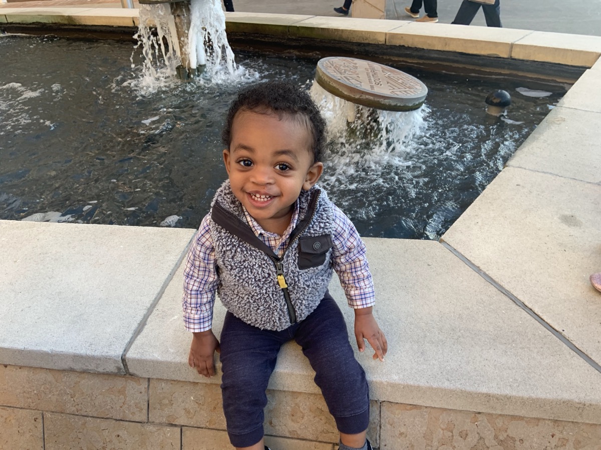 Yonas smiling brightly sitting on the edge of a water fountain outdoors, wearing a plaid button-down shirt, fleece vest, and dark jeans