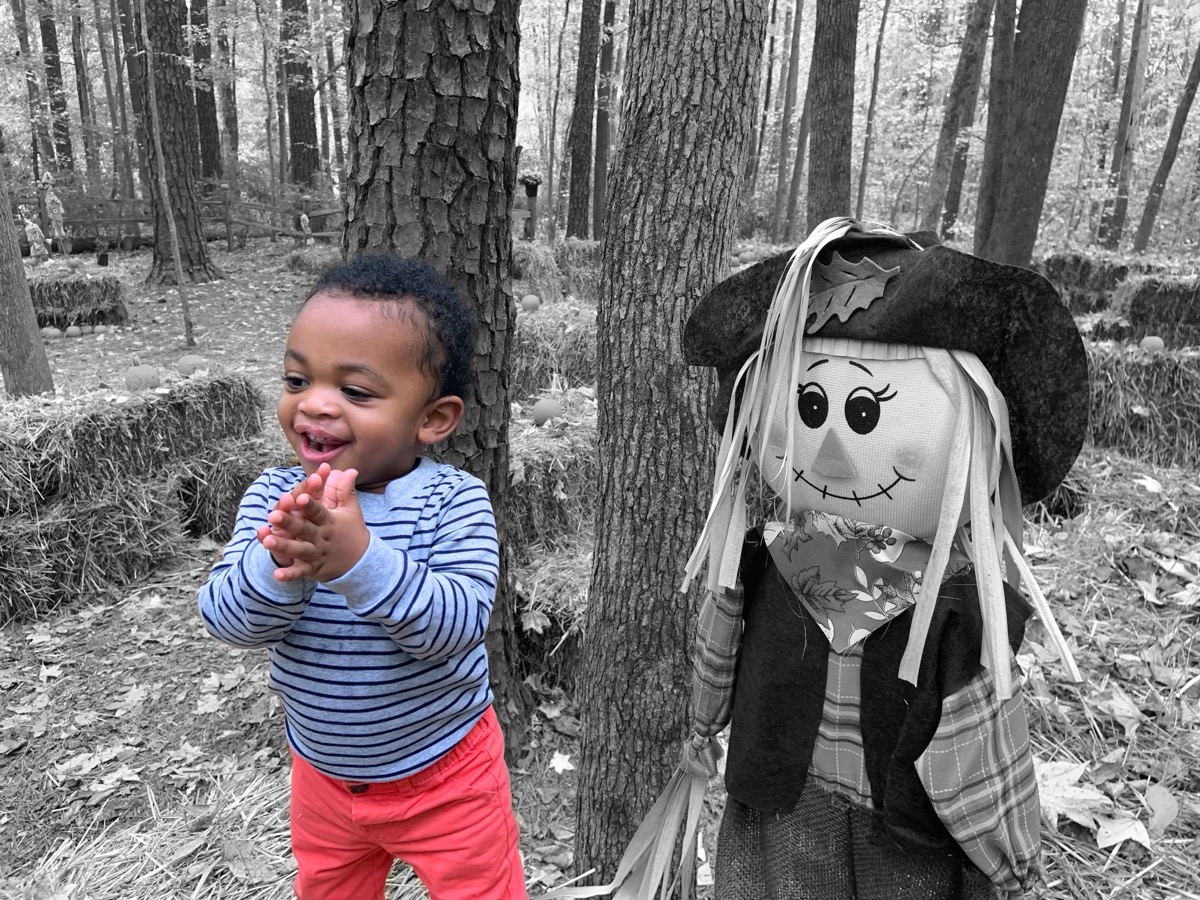 Yonas smiling and reaching forward next to a scarecrow at a fall pumpkin patch, wearing a striped long-sleeve shirt and bright red pants
