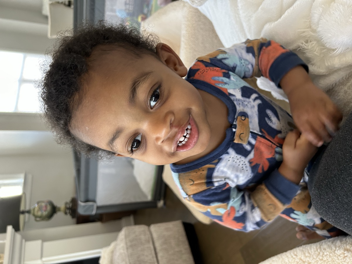 Yonas sitting on a couch in animal-print pajamas with curly hair, smiling sweetly at the camera in the living room