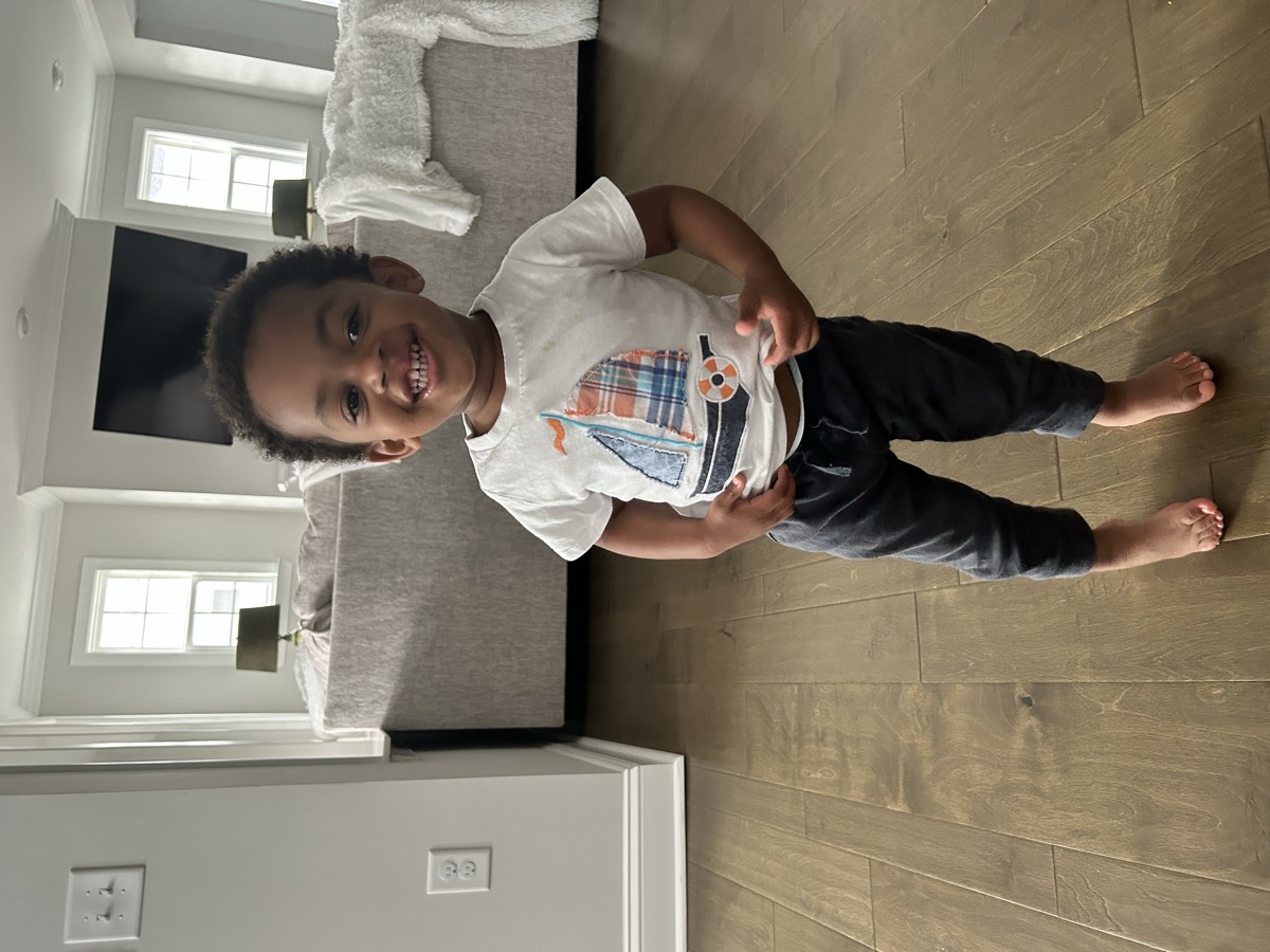 Yonas standing barefoot in the living room with a big grin, wearing a white sailboat graphic tee and dark joggers