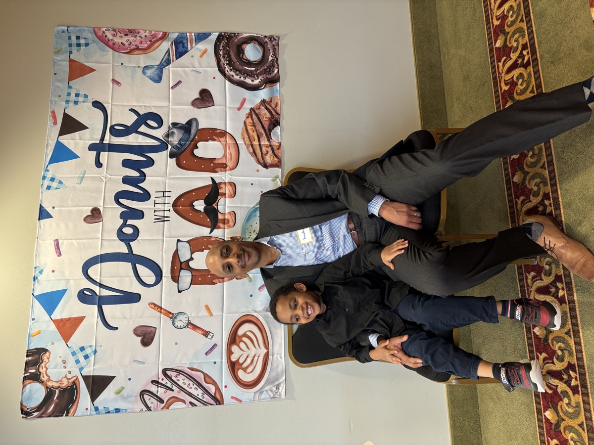 Yonas and his dad dressed up at a Donuts with Dad school event, both in dark suits, Yonas beaming on his dad's lap